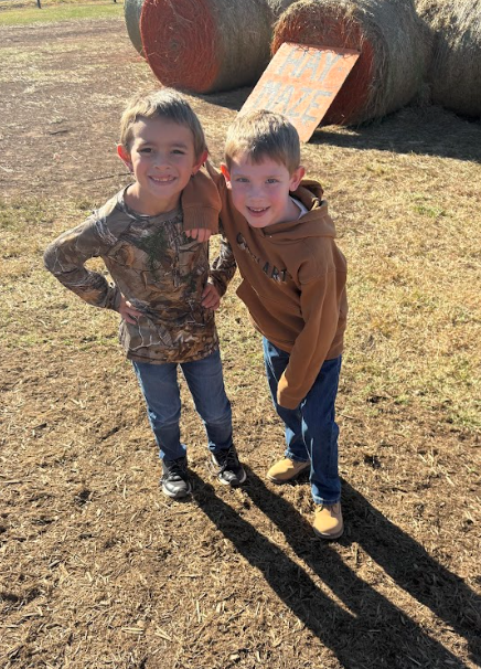 Two boys stand side by side outdoors, smiling with arms around each other near hay bales labeled “Hay Maze.”