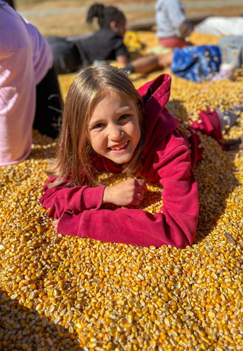 Photo 2: A smiling girl in a red hoodie lies on a pile of yellow corn kernels with sunlight shining on her face.