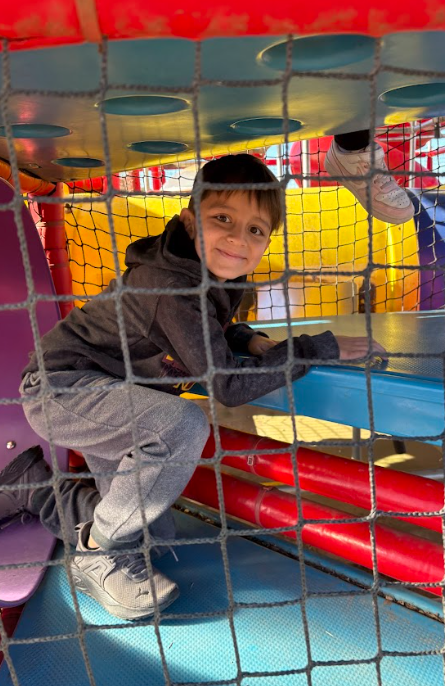 A child in a gray outfit smiles from inside a colorful climbing play structure behind a safety net.