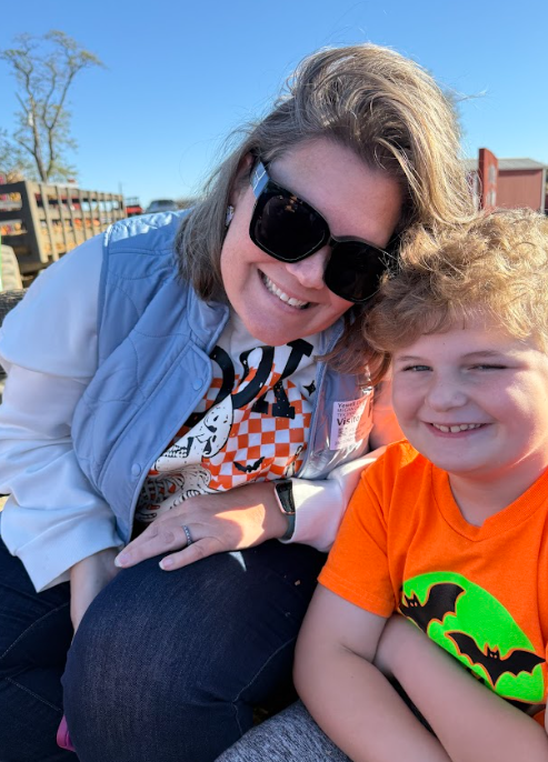 Photo 3: An adult wearing sunglasses and a light blue vest sits next to a smiling child in an orange Halloween-themed shirt during the field trip.