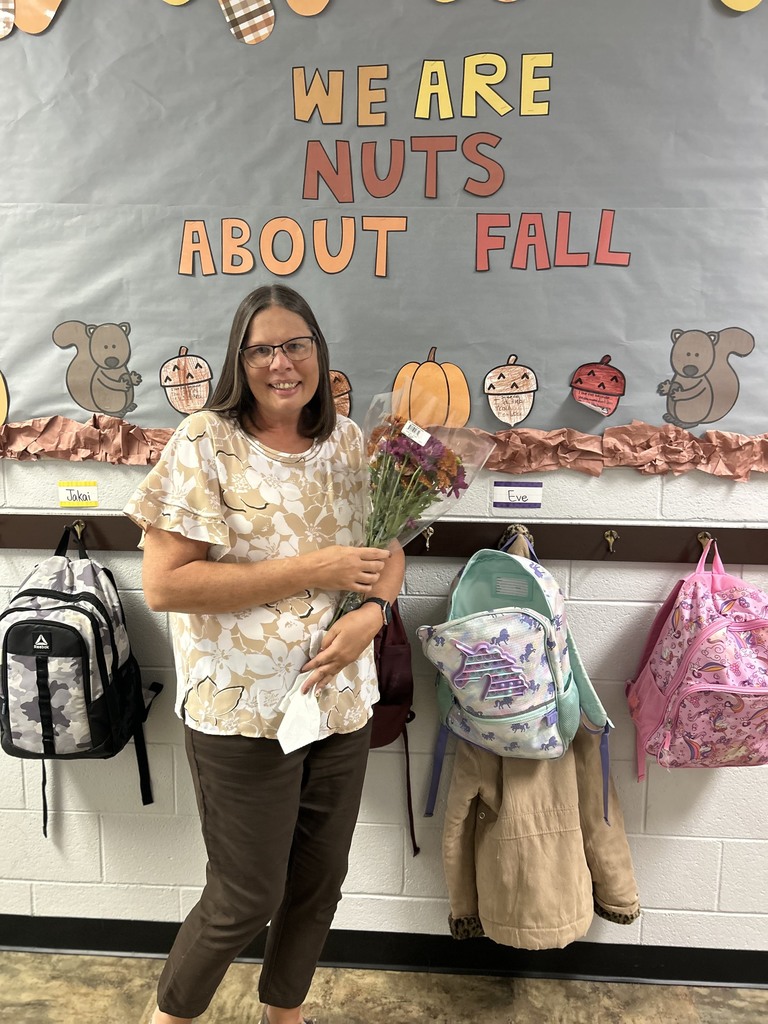 A smiling woman holding a bouquet of flowers stands in front of a classroom bulletin board that says “We Are Nuts About Fall.” The board is decorated with pumpkins, acorns, and squirrels. Backpacks hang on hooks along the wall behind her.