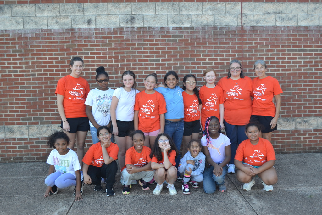 The Girls on the Run club pose in their orange shirts in front of a brick wall.  There are two rows of girls and three adults in the back row.