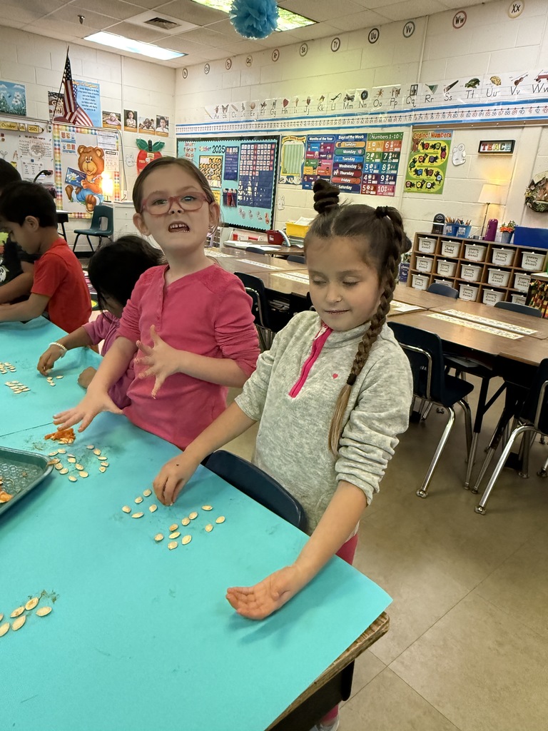 A photo of a smiling girl sitting at a table in the classroom, with other students and classroom materials visible in the background.