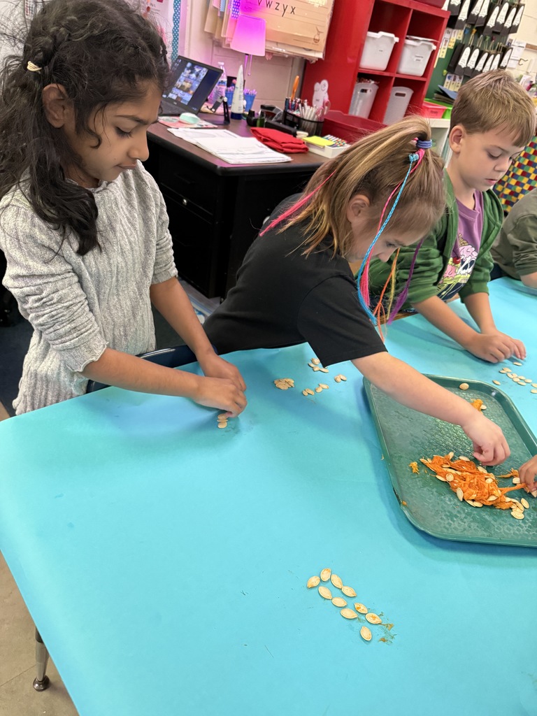 Several young children clustered around a table, actively involved in a classroom activity. Two girls in the foreground are looking down at a task in front of them.