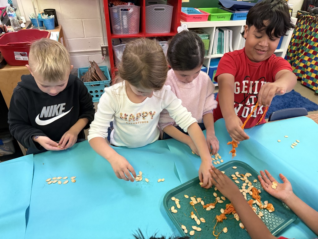 Multiple young students seated around a bright blue table, working on an activity together, with various expressions of focus and engagement.