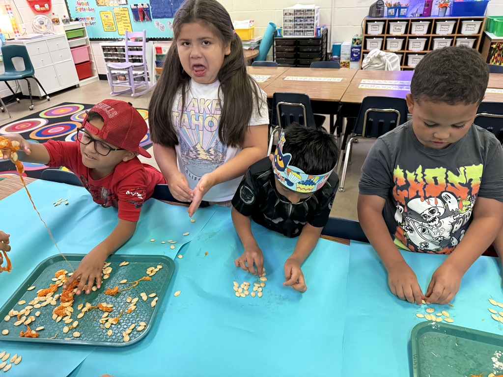 Two children, a boy and a girl, smiling and looking up from their task while sitting at a table with other classmates. They are in a brightly lit classroom setting.