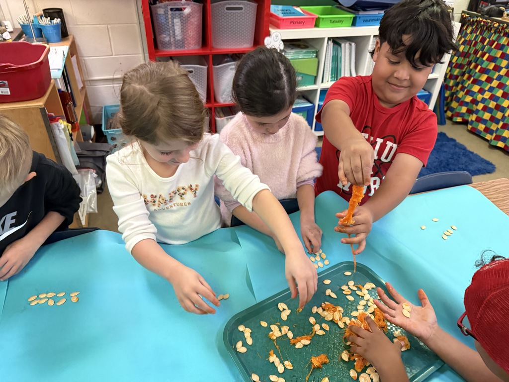 A group of students sitting at a blue table, with several children reaching toward the center of the table. They appear to be counting or sorting small items.
