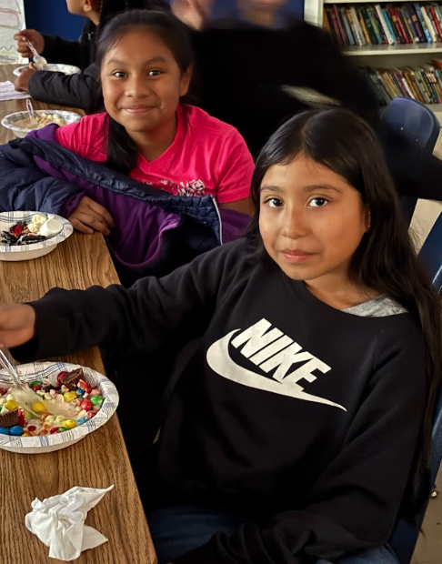Two 5th grade students sit at a classroom table smiling at the camera. One wears a pink shirt and the other a black Nike sweatshirt. Both have bowls of ice cream sundaes topped with colorful candy pieces in front of them.