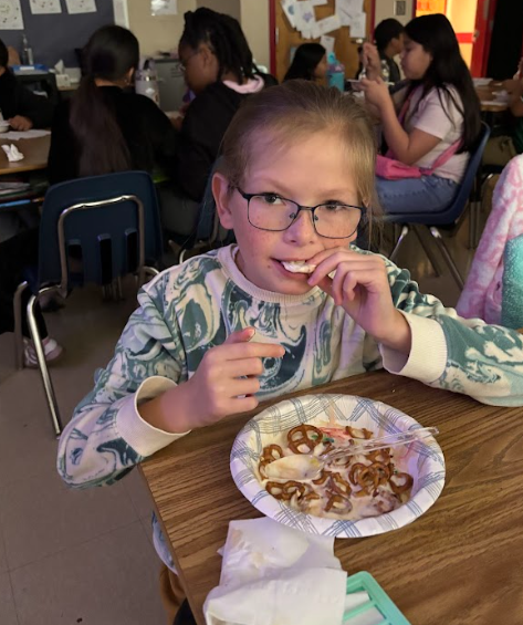 A student wearing glasses and a green-and-white patterned sweatshirt eats ice cream with pretzels on top. Other classmates can be seen seated at desks in the background.