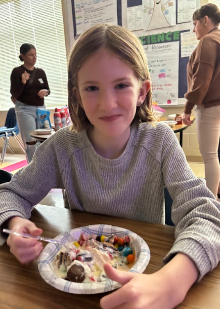 A student wearing a gray long-sleeve shirt smiles while holding a spoon over a bowl of ice cream with M&Ms and other toppings. Other students and a teacher are visible in the background.