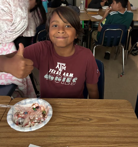A student wearing a maroon Texas A&M Aggies shirt gives a thumbs up while sitting at a desk with a bowl of ice cream covered in toppings. Other students are seated in the background.
