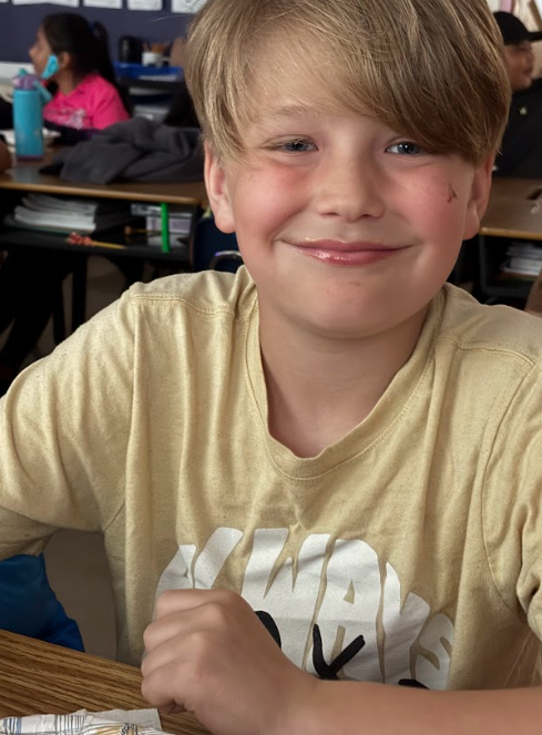 A smiling student in a yellow T-shirt sits at a desk with a paper plate of ice cream nearby. Other students are seated behind him.