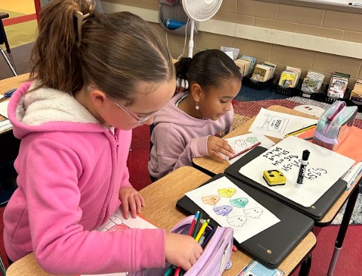 Two students sit at their desks working on Halloween-themed math worksheets. Both are wearing pink hoodies and focusing on coloring and solving problems using pencils and markers.