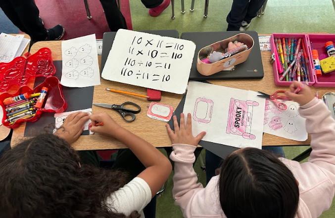 Two students color Halloween-themed math sheets at a table covered with art supplies, scissors, and markers. A whiteboard nearby shows multiplication equations like “10 x 10 = 100.”