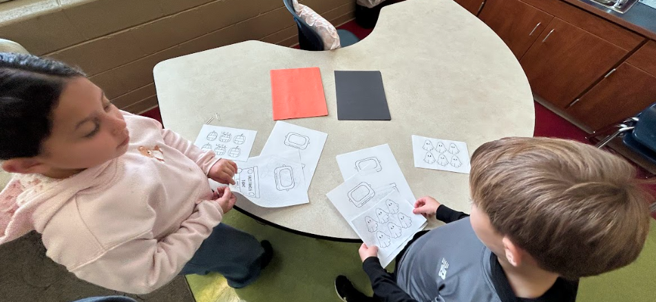 Two students stand at a kidney-shaped table looking over printed pages with ghost and pumpkin math designs as they prepare their projects.