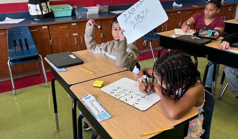 Two students work on math problems at their desks. One holds up a whiteboard showing a completed multiplication equation, while another writes her answers carefully on her own board.