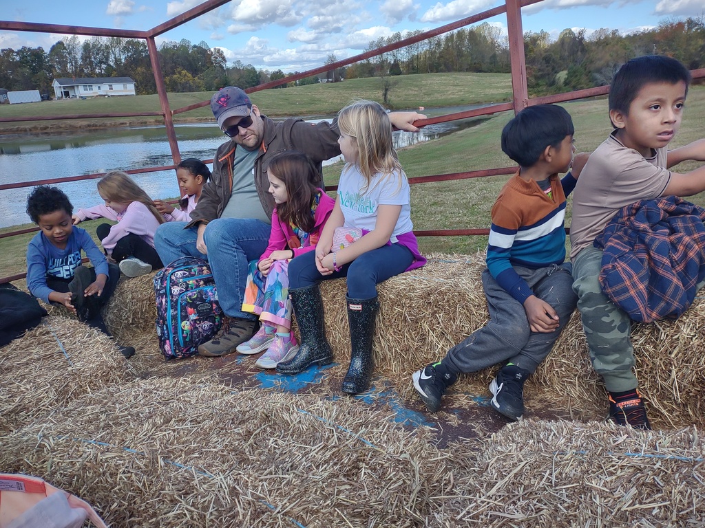 A group of children and an adult man sitting on hay bales inside a metal-framed wagon on a sunny day. A pond or lake and grassy field are visible in the background.