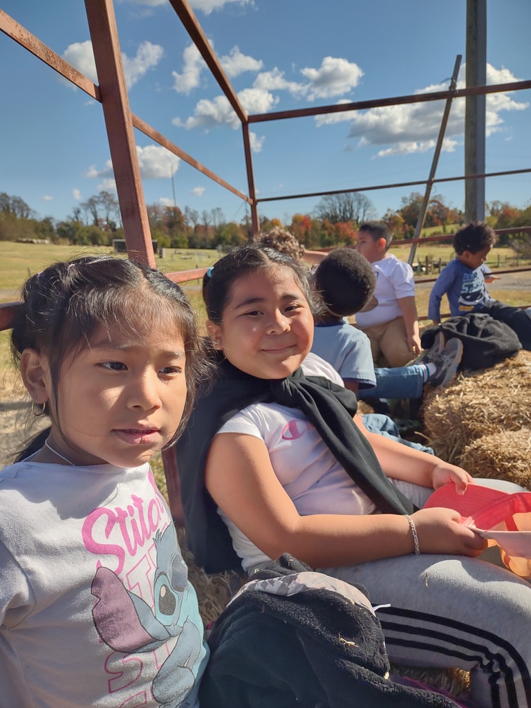 Two young girls sitting on a hay bale inside an open-air wagon or trailer. The girl on the right is smiling at the camera, and other children are visible in the background, also sitting on hay.
