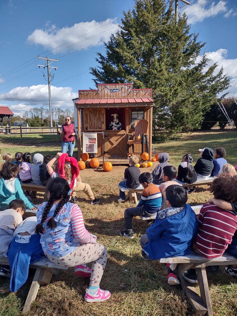 A group of elementary-aged children sitting on wooden benches outdoors, attentively watching an adult (a woman in a red plaid shirt) standing near a small wooden puppet stage. Several pumpkins are placed around the stage.