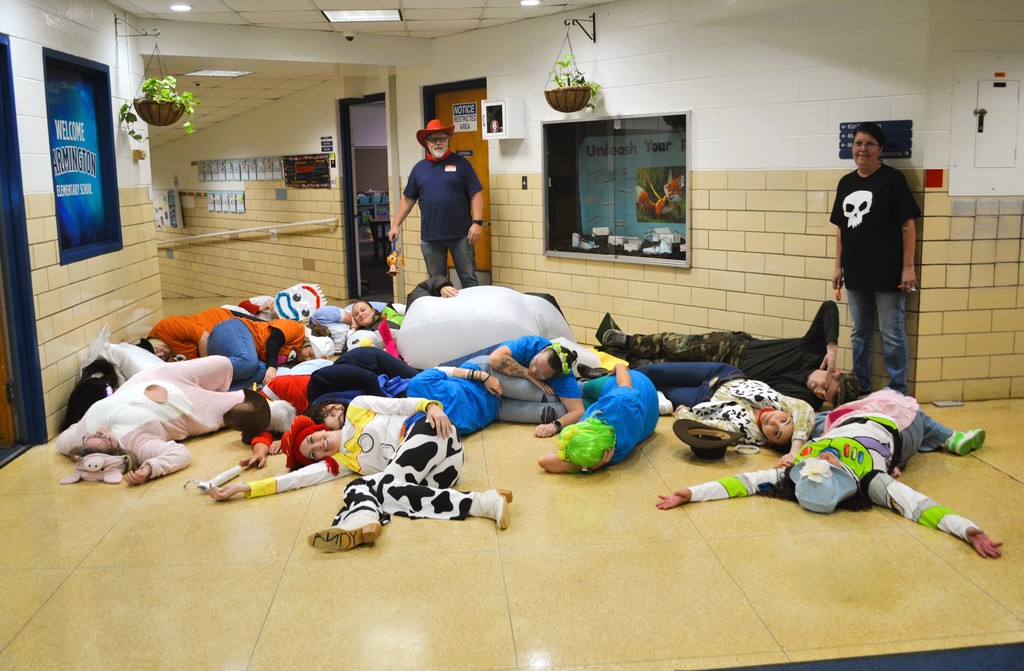  An image of a group of people, many dressed in Toy Story character costumes, lying motionless on the floor of a school hallway, mimicking the scene where the toys "go limp" when their owner, Andy, enters the room.  The scene includes:  A person dressed as Woody (in the foreground, lying face-up with a red hat and cow-print chaps).  A person dressed as Jessie (lying next to Woody, in a white shirt and red pigtails).  A person dressed as Buzz Lightyear (lying on their stomach, visible on the right with a green and white costume).  Other individuals are in various poses and costumes, some resembling other Toy Story characters or generic clothing.  Two adults are standing on the periphery, looking at the group on the floor. One is a man in a dark blue shirt and a cowboy hat, and the other is a woman in a black t-shirt with a white skull design.  The setting appears to be a school hallway, with a sign reading "WELCOME TO Farmington ELEMENTARY SCHOOL" visible on the left wall.  The walls have a two-tone pattern of white and beige brick.  Potted plants hang from the ceiling.  The floor is light-colored tile.  This image captures a playful group activity, likely a costumed event at the school.