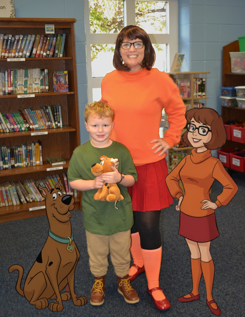 A woman dressed as Velma from Scooby-Doo (wearing an orange turtleneck, red pleated skirt, black tights, orange knee socks, and red shoes) stands next to a smiling young boy. The woman is wearing glasses and has her hands on her hips. The boy wears a green t-shirt, tan pants, and brown boots, and holds a small brown stuffed animal. They are in a library or media center, with a wooden bookshelf full of books to their left and a bright window behind them. Two large cartoon cutouts of Scooby-Doo (sitting on the left) and Velma (standing on the right) are placed on the carpeted floor next to them.