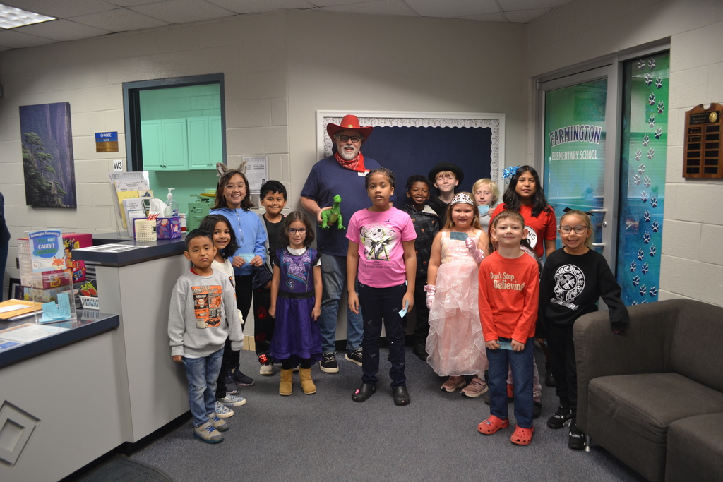 A dozen smiling students in a variety of outfits and costumes pose with the principal (dressed as Andy from Toy Story) in the main office of the school.