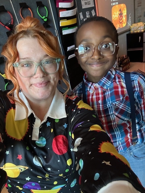 A woman and child smiling. The woman wears glasses and a space-themed shirt. The child is in glasses, a plaid shirt, and suspenders. Classroom setting.