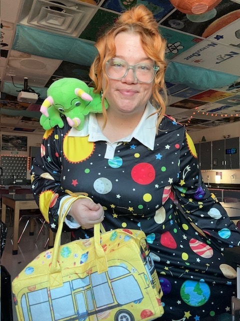 A person with glasses smiles brightly indoors, wearing a colorful, planet-patterned dress. A green plush dinosaur rests on their shoulder, and they hold a yellow bus-patterned bag. The setting has a playful and cheerful vibe.