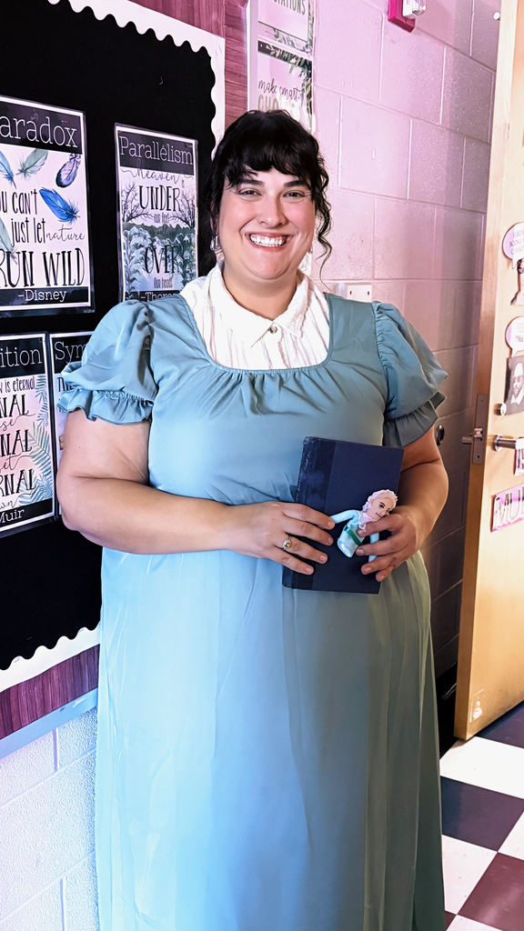 A woman in a light blue dress and white collar smiles, holding a blue book with a small doll. She stands in a classroom with educational posters.