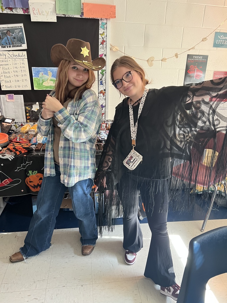 Two people are dressed for Halloween. One wears a cowboy hat and plaid shirt, the other a fringed black outfit. A decorated table and posters are in the background.