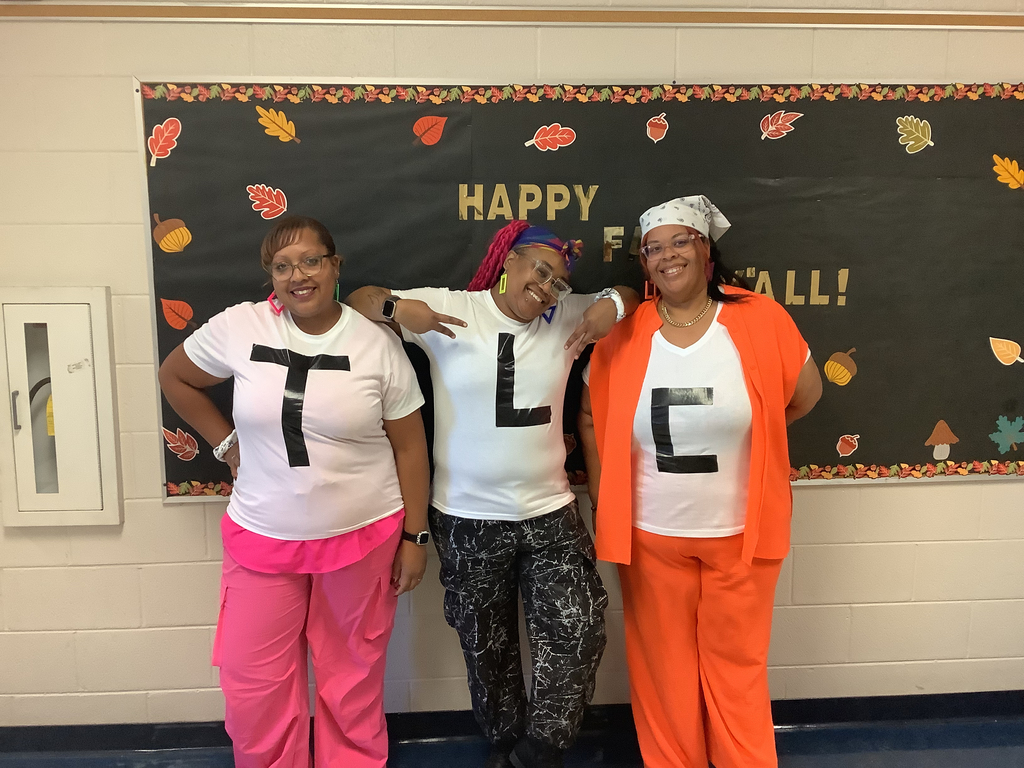 Three women in front of a fall-themed bulletin board wear white shirts with "T," "L," and "C" taped on them, smiling and posing joyfully.
