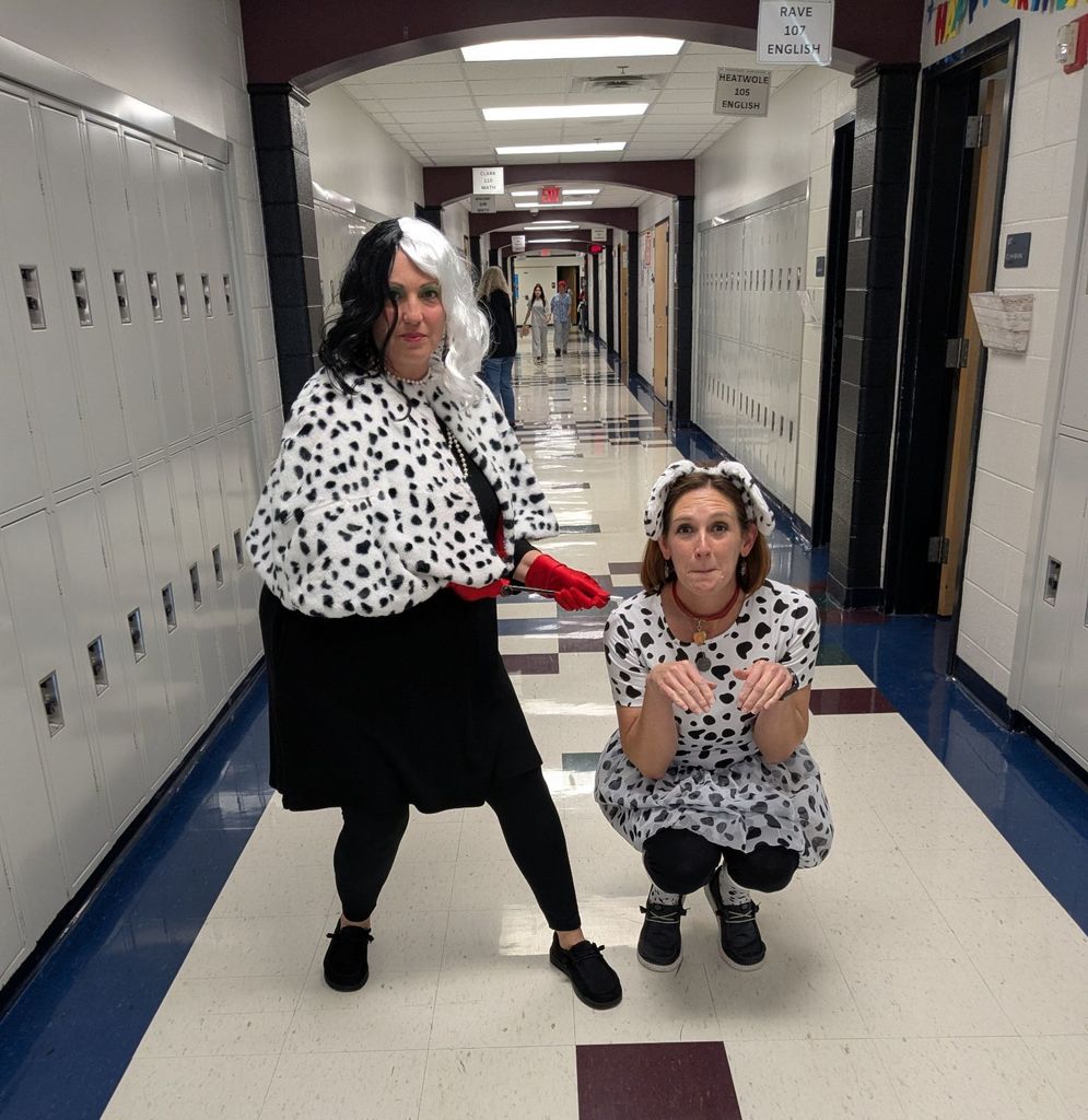 Two people in a school hallway wear Dalmatian-inspired costumes. One stands in a black and white wig and cape, while the other crouches in a matching dress. Fun atmosphere.