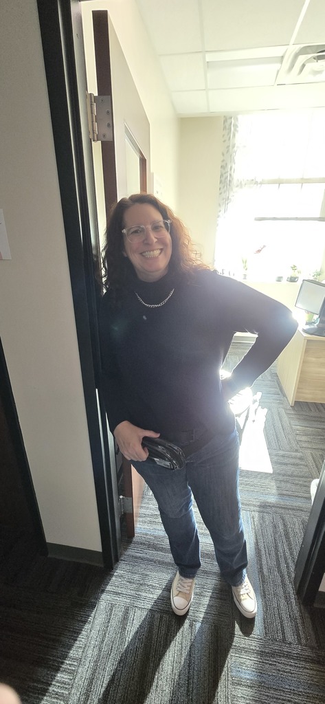 A woman with curly hair and glasses stands smiling in a sunlit office. She's wearing a black sweater, jeans, and sneakers, exuding a friendly vibe.