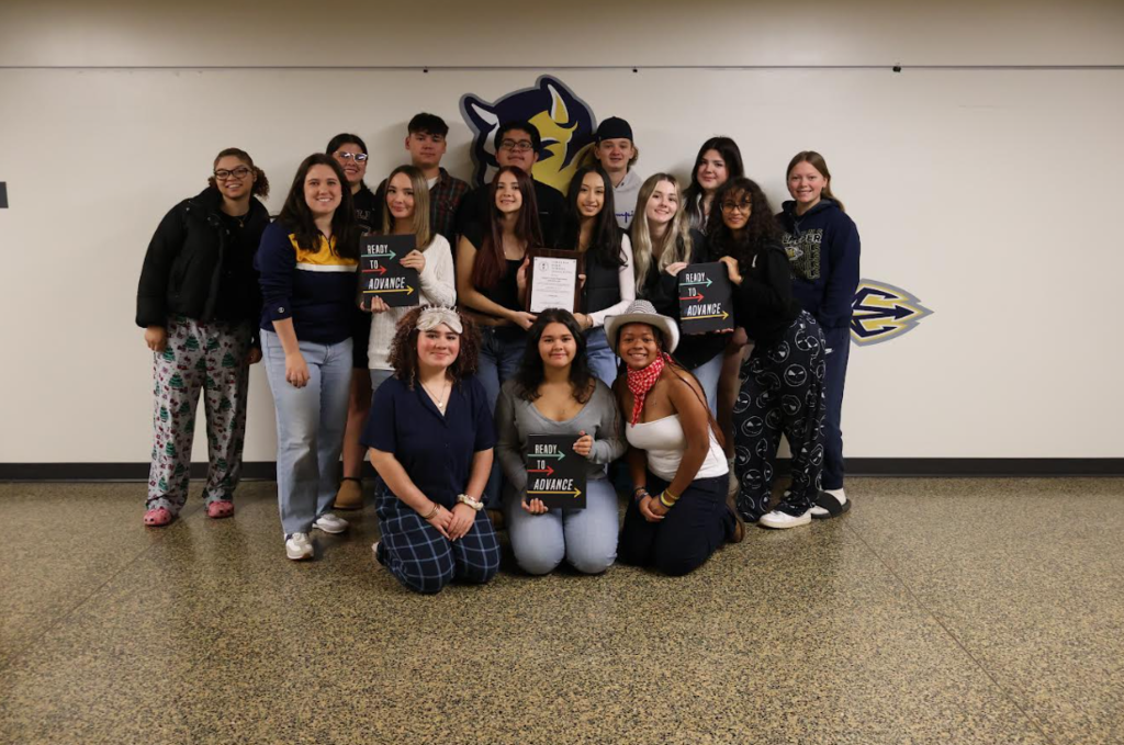 Mrs. Rogers and 15 yearbook students stand in front of the Culpeper County High School logo on a wall. The person in the middle holds a framed certificate, while the two on either side hold “Ready to Advance” signs. All are smiling and dressed in Culpeper County High School apparel, celebrating VHSL Trophy Class yearbook recognition.