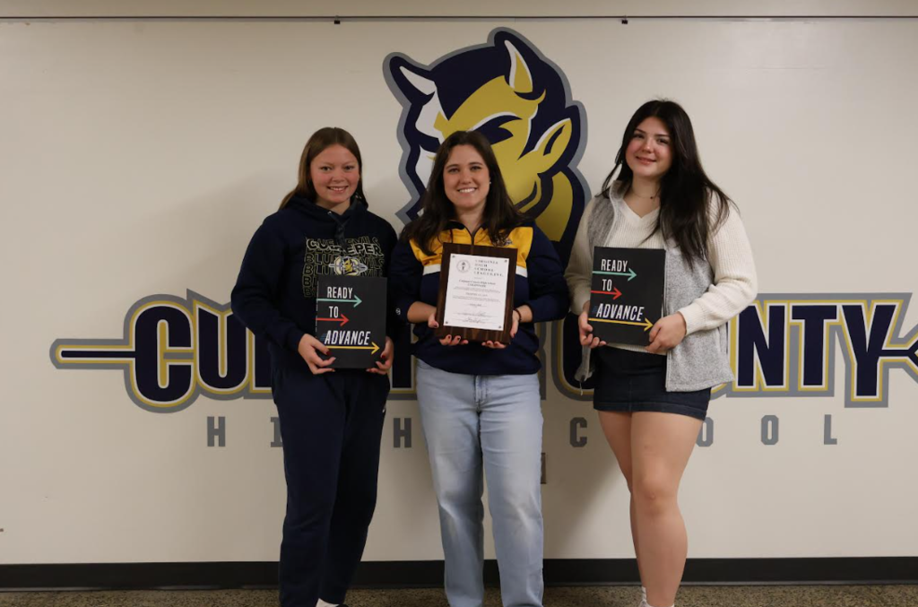 Mrs. Rogers and 2 students stand in front of the Culpeper County High School logo on a wall. The person in the middle holds a framed certificate, while the two on either side hold “Ready to Advance” signs. All are smiling and dressed in Culpeper County High School apparel, celebrating VHSL Trophy Class yearbookrecognition.