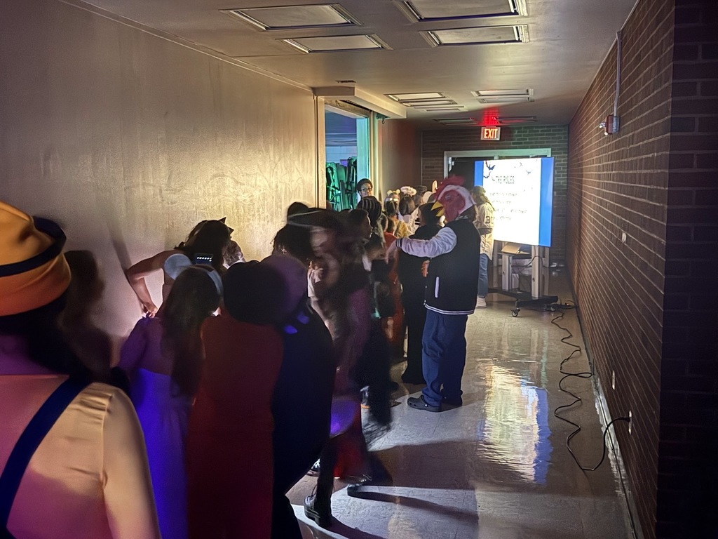 A group of people, many in costumes, are walking down a narrow hallway with a brick wall on the right. An exit sign and a projected screen are visible at the end of the hall.