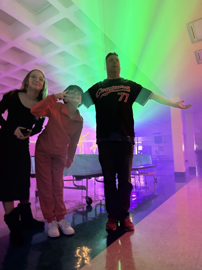 A photograph taken indoors under dramatic, vibrant purple and green stage lighting. A person is standing center, facing away from the camera, wearing a dark baseball-style jersey with 'Cougars 77' stitched across the back. They have their arms stretched out to the sides. In the middle ground, there is a low metal cart, possibly a gurney or hospital bed, with a delicate, abstract, golden-colored sculpture on it. Other people and a wall with two levels of windows are visible in the background, mostly obscured by the dark, smoky, and colorful atmosphere of what appears to be an event or performance.