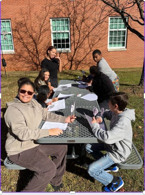 A group of children sits at an outdoor metal picnic table, smiling and working on crafts under a sunny sky. A brick building with windows is in the background.