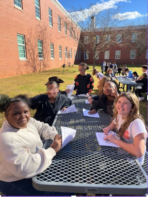 A group of students sits at outdoor tables with worksheets, enjoying a sunny day. The scene conveys a cheerful, collaborative atmosphere.