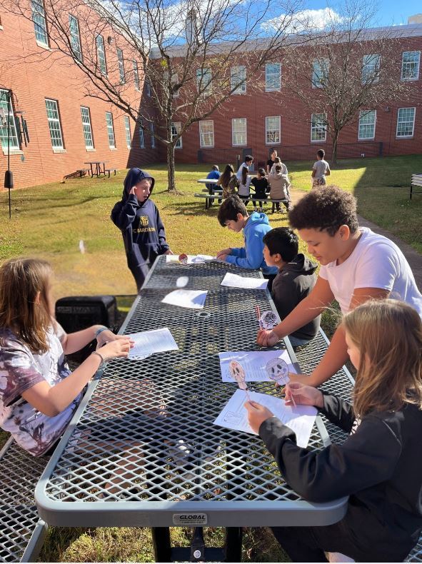 Students gather around outdoor picnic tables, working on papers under a clear blue sky. The scene conveys focus and collaboration in a school courtyard.