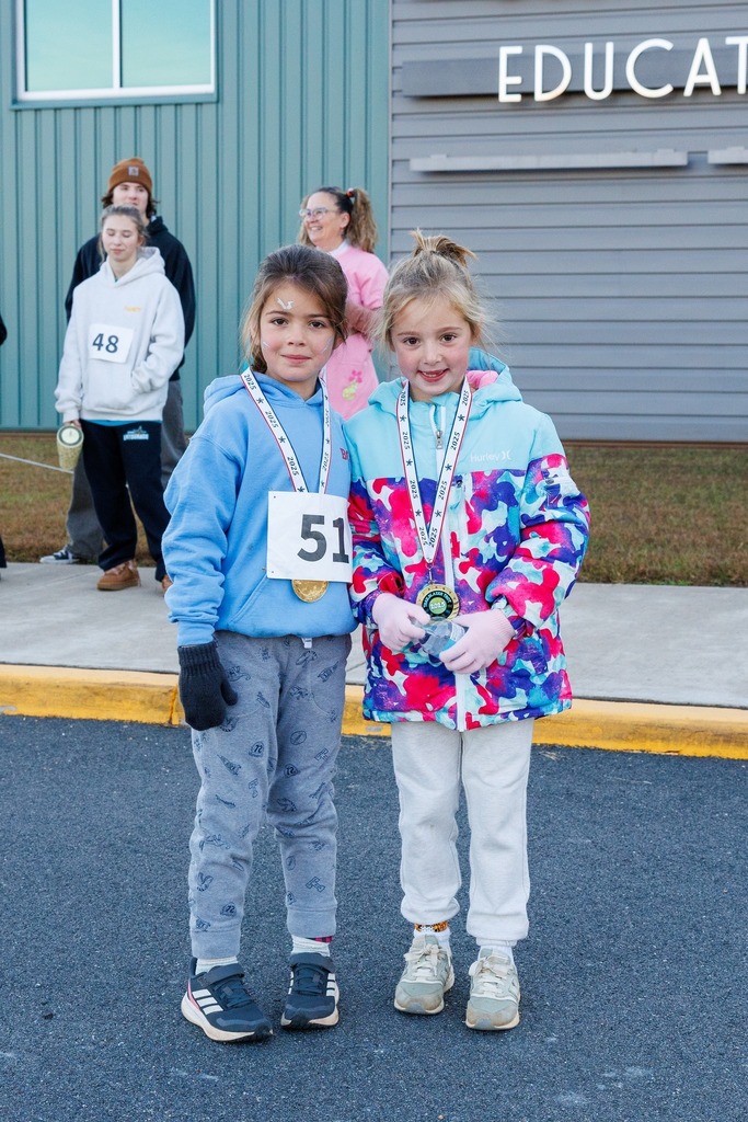 Two young girls wearing medals pose together after completing their run, smiling proudly at the finish area.