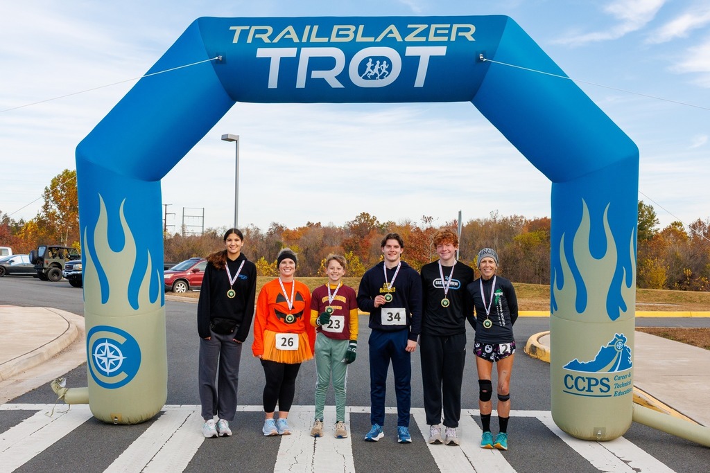 A group of Trailblazer Trot winners stand proudly under the blue finish line arch, each wearing medals and smiling for the camera.