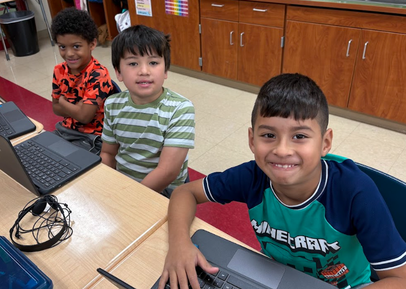 Three second-grade students sit side by side at their desks using Chromebooks. They smile at the camera—one in an orange shirt, one in a green striped shirt, and one in a blue Minecraft shirt—with classroom cabinets in the background.