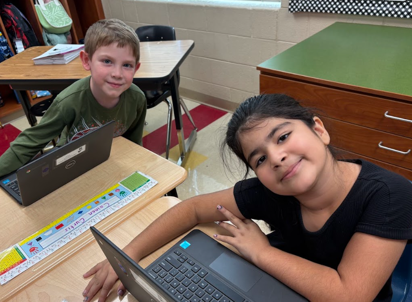 Two second-grade students smile at their desks while working on Chromebooks during an Internet safety lesson. The boy wears a green long-sleeve shirt, and the girl wears a black top, both seated in a bright classroom.