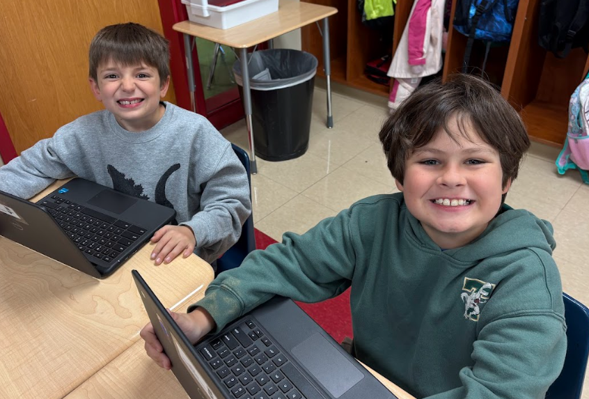 Two second-grade boys smile at their desks with Chromebooks open in front of them. One wears a gray sweatshirt and the other a green hoodie.