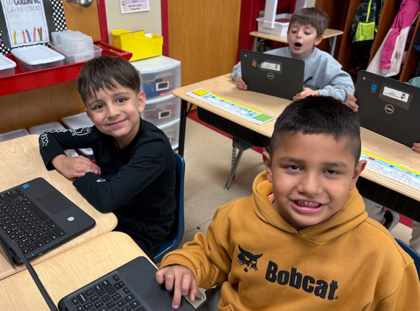 Two second-grade boys work on Chromebooks and smile at the camera. The student in front wears a tan hoodie that says “Bobcat,” and the student beside him wears a black long-sleeve shirt.