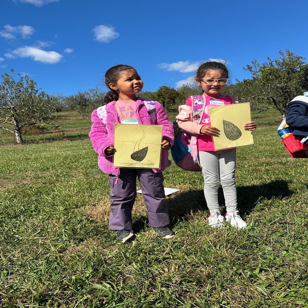 Two young girls stand side by side outdoors in a grassy field holding large paper drawings of seeds and sprouts. The sky is bright blue with apple trees in the background.