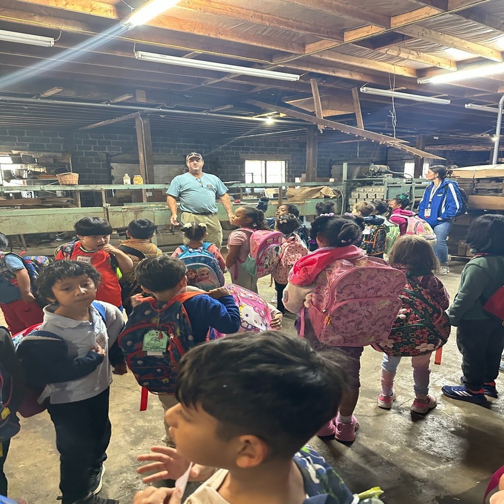 A group of elementary students listen attentively to a farmer speaking inside a rustic barn. The children are wearing backpacks and jackets, gathered in front of wooden farm equipment.