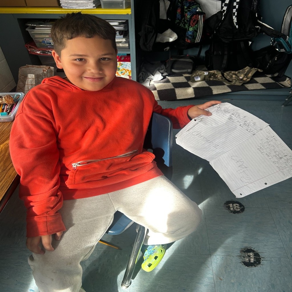 A student in a red hoodie sits at a classroom desk holding up handwritten story pages with a smile.
