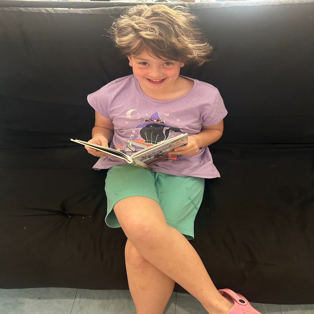 A smiling student in a purple shirt and teal shorts sits on a black couch reading a book.
